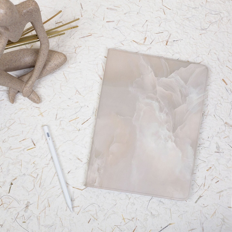 May include: A rectangular tablet case featuring a soft, beige and white marbled pattern with a smooth finish. Positioned next to the case is a slim, white digital stylus. The case has rounded corners and a clean, flat surface design.