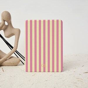 May include: A rectangular electronic reader cover featuring a vertical striped pattern in alternating shades of light pink and pale yellow. The lower center of the cover displays the gold-colored initials CL in a simple, clean serif typeface.