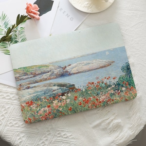 Op de afbeelding: Een rechthoekige laptoptas met een schilderij van een kustlandschap. Het kunstwerk toont een rotsachtige kustlijn met rode en witte bloemen, een blauwe zee en een lichtblauwe lucht. De hoes ligt op een witte, getextureerde ondergrond.
