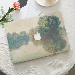 Peut inclure: Une coque d'ordinateur portable avec un motif de paysage impressionniste dans les tons de vert, bleu et beige. Elle présente une découpe du logo Apple blanc. La coque est posée sur une surface blanche, avec une fleur et de la verdure.