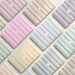 May include: A collection of laptop cases featuring various pastel-colored stripes. Each case displays a cutout for the Apple logo. The colors include soft pink, light blue, lavender, mint green, and pale yellow, all arranged in a repeating pattern.