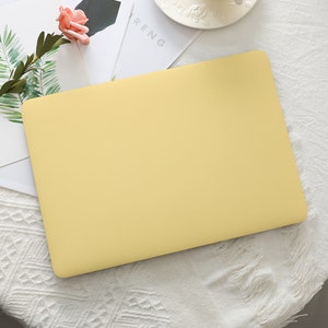 Può includere: Un computer portatile giallo pallido su una superficie bianca. Il computer portatile è chiuso e ha una finitura liscia e opaca. Una rosa rosa e foglie verdi sono visibili sullo sfondo, insieme a una tovaglia bianca.