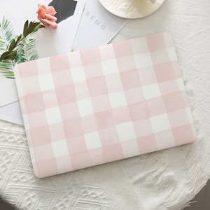 May include: A pink and white gingham laptop sleeve. The sleeve features a watercolour effect with soft, blended edges. The pattern is a classic plaid design. The rectangular sleeve appears to be made of a soft material, ideal for protecting a laptop.