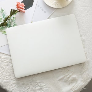 May include: A white laptop computer on a white tablecloth. A pink rose and green leaves are on a white card next to the laptop. A white teacup and saucer are in the background.