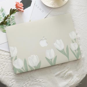 Könnte beinhalten: Eine hellbeige Laptop-Hülle mit einem Blumendesign aus weißen Tulpen mit grünen Stielen und Blättern. Die Hülle hat einen Ausschnitt für das Apple-Logo. Das Wort "RENG" ist auf einem Papier zu sehen.