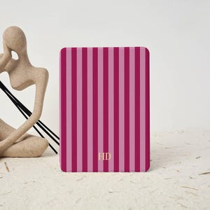 May include: A rectangular e-reader cover featuring a vertical striped pattern in alternating shades of dark magenta and light pink. The letters HD are printed in a small, gold-colored font centered near the bottom edge of the front cover.