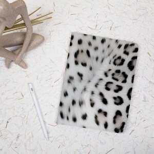 May include: A rectangular notebook cover featuring a white fur pattern with black leopard spots. The cover has a soft, textured appearance and is positioned next to a white stylus on a light, speckled surface.