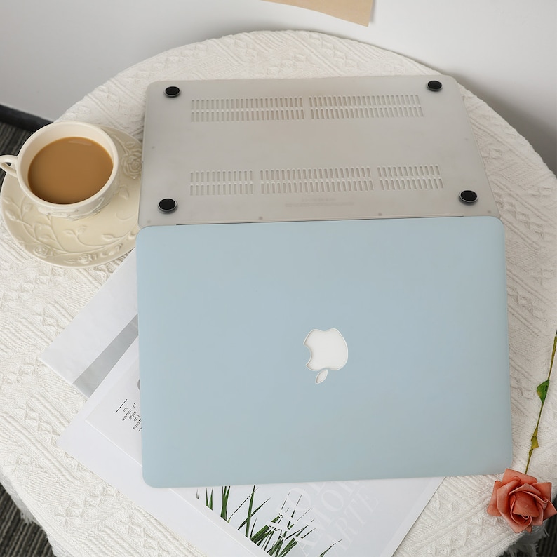 May include: A light blue laptop with a white Apple logo is displayed open, with a clear protective cover on top. The laptop is on a white table with a cup of coffee and a rose.