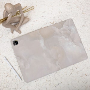 May include: A rectangular tablet case featuring a light beige and white marbled pattern with a smooth finish. The case includes a precise cutout for a dual-lens camera system. A slim white stylus is positioned diagonally next to the tablet case.