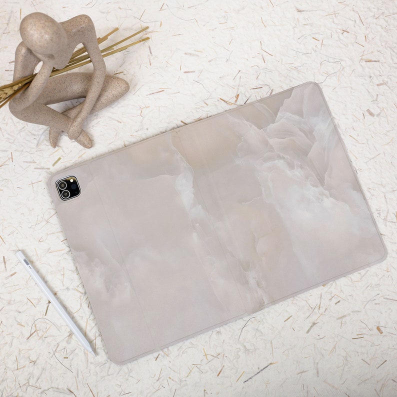 May include: A rectangular tablet case featuring a light beige and white marbled pattern with a smooth finish. The case includes a precise cutout for a dual-lens camera system. A slim white stylus is positioned diagonally next to the tablet case.