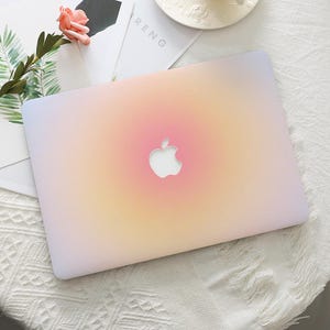 Pode incluir: Um laptop com um design gradiente em tons de rosa, amarelo e lavanda. O logotipo da Apple é visível no centro. O laptop está em uma superfície branca.