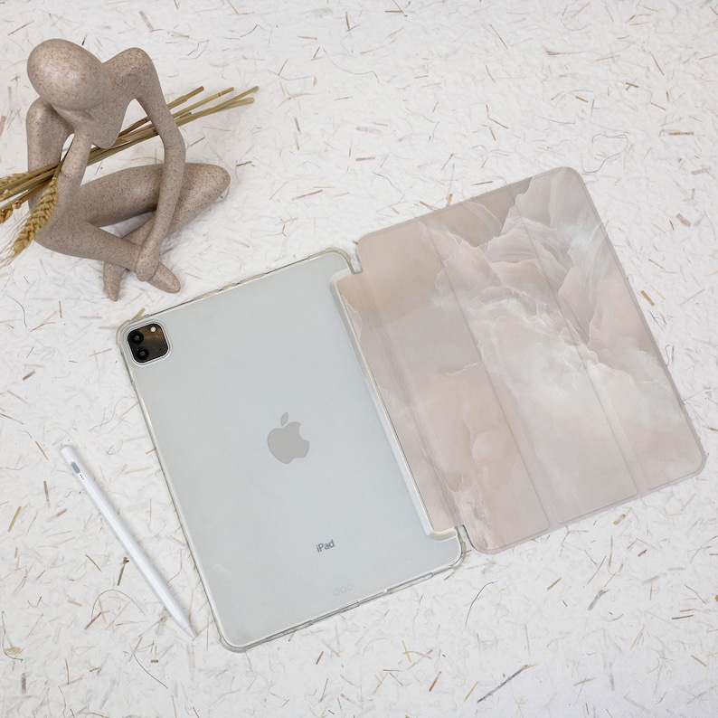 May include: A silver iPad with a clear protective case and a white stylus. The device is paired with a folding cover featuring a soft, marbled beige and white pattern. A small, abstract, light brown human-like figurine is positioned nearby.