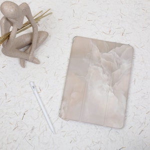 May include: A rectangular tablet case featuring a beige and white marbled pattern with a tri-fold design. Beside the case lies a slim white stylus pen. A beige abstract human-shaped figurine holding thin wooden sticks is positioned nearby.