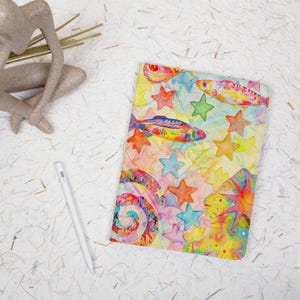 May include: A rectangular notebook cover featuring a vibrant watercolor illustration of colorful fish, swirling patterns, and various stars in shades of yellow, orange, pink, blue, and green. The design is set against a light, soft-toned background.