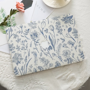Op de afbeelding: Een witte laptophoes met een blauw bloemenpatroon. Het ontwerp bevat verschillende botanische illustraties van bloemen en bladeren. De hoes heeft een uitsparing voor het Apple-logo en de algehele esthetiek doet denken aan een botanische print.