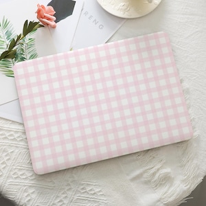 May include: A pink and white gingham laptop case. The case is rectangular and features a classic checkered pattern. The case is shown on a white surface with a flower and greenery in the background.