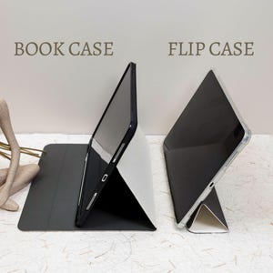 May include: A black book case and a flip case for a tablet are displayed side by side. The book case features a dark cover and a stand, while the flip case has a clear shell and a white stand. Both cases are shown in a propped-up position.