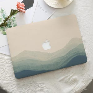 May include: A laptop case with a mountain landscape design. The case features a beige top with a cut-out Apple logo and a watercolour-style depiction of layered blue and green mountains across the bottom.