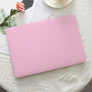 Può includere: Una custodia per laptop rosa chiaro. La custodia rettangolare ha angoli arrotondati. È appoggiata su una superficie bianca, con una composizione floreale e una rivista sullo sfondo.