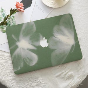 Puede incluir: Una funda verde para portátil con un diseño floral blanco. La funda tiene un logotipo de Apple blanco en el centro. La palabra "RENG" es visible en un papel blanco en el fondo.