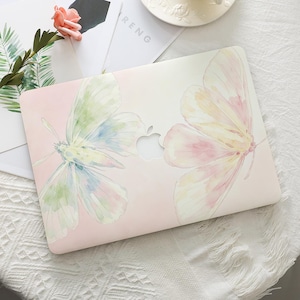 May include: A laptop hard shell case featuring a soft watercolor illustration of two butterflies with pastel pink, green, and yellow wings. The design is set against a pale pink gradient background and includes a central cutout for the Apple logo.