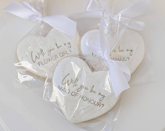 May include: Three heart-shaped biscuits with white icing and gold lettering, each individually wrapped in clear plastic bags tied with white satin ribbons. The text on the biscuits reads Will you be my FLOWER GIRL?, Will you be my BRIDESMAID?, and Will you be my MAID OF HONOUR?.