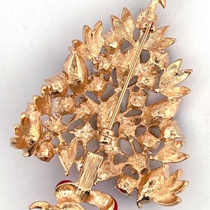 May include: Gold-coloured Christmas tree brooch with a star on top and a red ribbon. The brooch features textured leaves and a pin closure. A festive accessory for the holiday season, perfect for a coat or scarf.