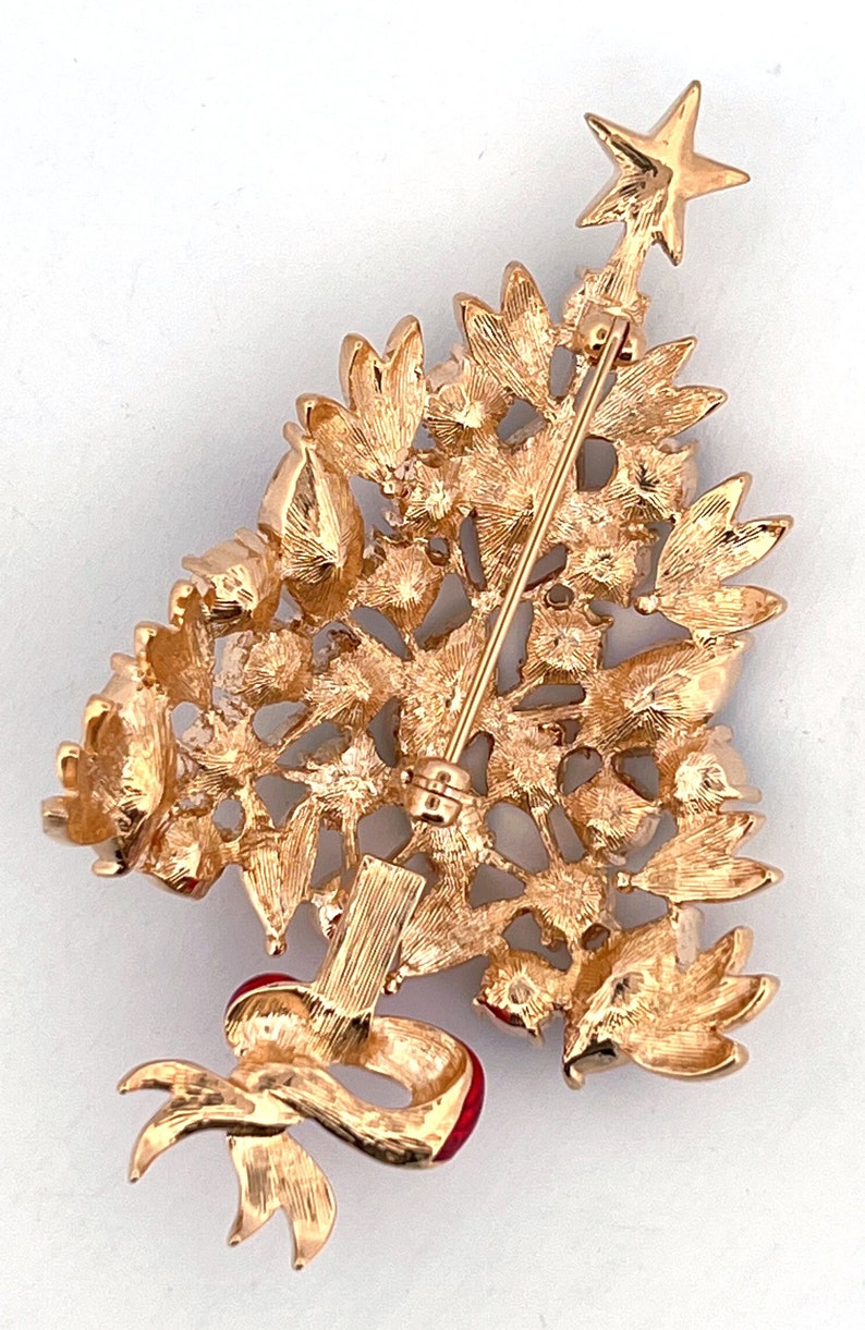 May include: Gold-coloured Christmas tree brooch with a star on top and a red ribbon. The brooch features textured leaves and a pin closure. A festive accessory for the holiday season, perfect for a coat or scarf.