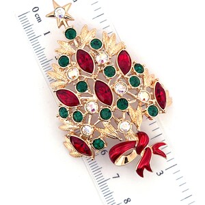 May include: Gold-toned Christmas tree brooch with red, green, and clear crystal accents. The brooch features a star at the top and a red bow at the base. The brooch is approximately 5 cm in length.