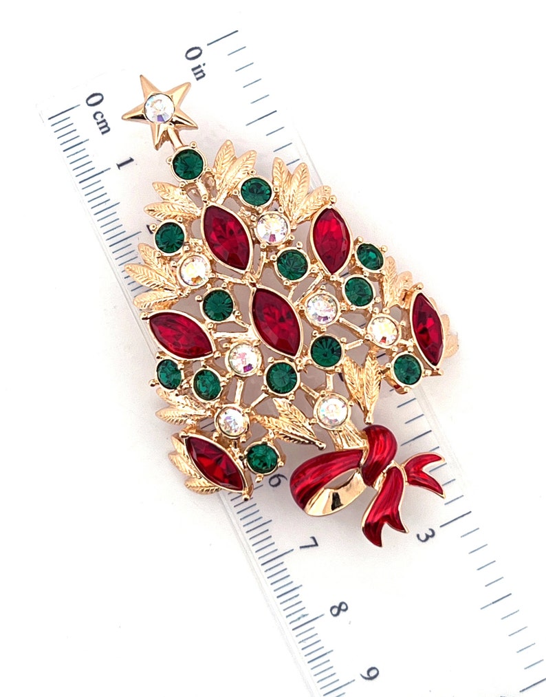 May include: Gold-toned Christmas tree brooch with red, green, and clear crystal accents. The brooch features a star at the top and a red bow at the base. The brooch is approximately 5 cm in length.