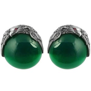 May include: A pair of silver earrings with green gemstone cabochons and a detailed eagle design. The eagle's eyes are set with small red gemstones.