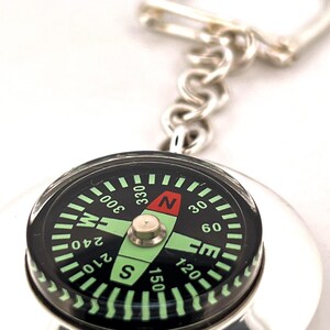 May include: A silver compass keyring with a black dial featuring green and white markings. The compass needle points north, indicated by a red arrow. The keyring has a silver chain and clasp.