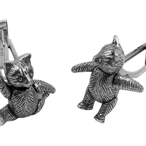 May include: A pair of silver cufflinks featuring a detailed teddy bear design. Each cufflink has a textured surface and a silver clasp.