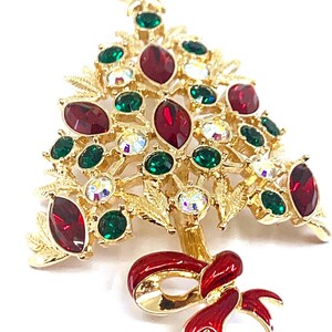 May include: Gold-coloured Christmas tree brooch with red, green, and clear gemstone accents. The brooch features a red enamel bow at the base and a star at the top. A festive holiday accessory.