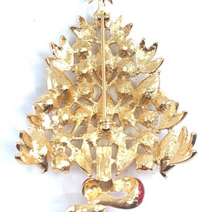 May include: Gold-coloured Christmas tree brooch with a star on top and a red bow at the base. The brooch features intricate detailing of leaves and ornaments, ideal for festive wear.