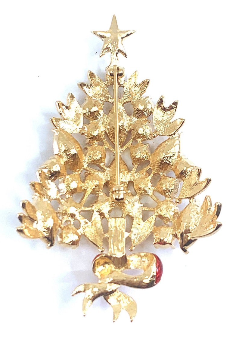May include: Gold-coloured Christmas tree brooch with a star on top and a red bow at the base. The brooch features intricate detailing of leaves and ornaments, ideal for festive wear.