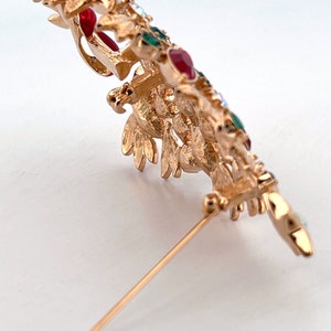 May include: Gold-coloured metal brooch in a bird design, embellished with red, green, and clear gemstones. The brooch has detailed features and a pin fastening. A decorative accessory for jackets, scarves, or hats.