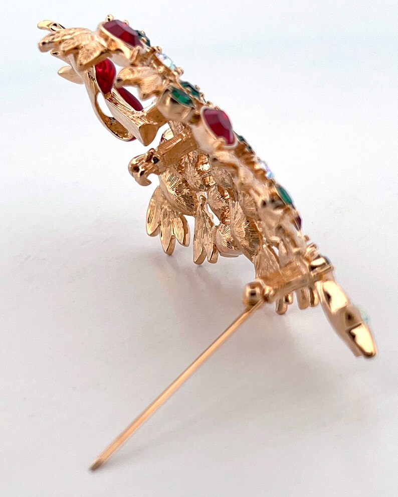 May include: Gold-coloured metal brooch in a bird design, embellished with red, green, and clear gemstones. The brooch has detailed features and a pin fastening. A decorative accessory for jackets, scarves, or hats.