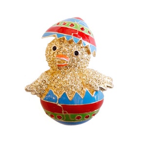 May include: A gold-tone chick hatching from a colorful Easter egg. The egg is decorated with red, blue, and green stripes. The chick is covered in sparkling rhinestones.
