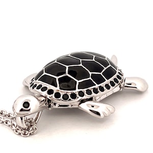 Könnte beinhalten: Eine silberne Schildkröten-Anhängerkette mit einer schwarzen Emaille-Schale und einer Reihe schwarzer Edelsteine ​​am Rand. Die Schildkröte befindet sich an einer silbernen Kette.