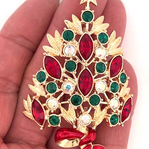 May include: A gold-coloured Christmas tree brooch with red, green, and clear crystal accents. The brooch features a star on top and a red ribbon at the base. The brooch is approximately 6.35 cm tall.