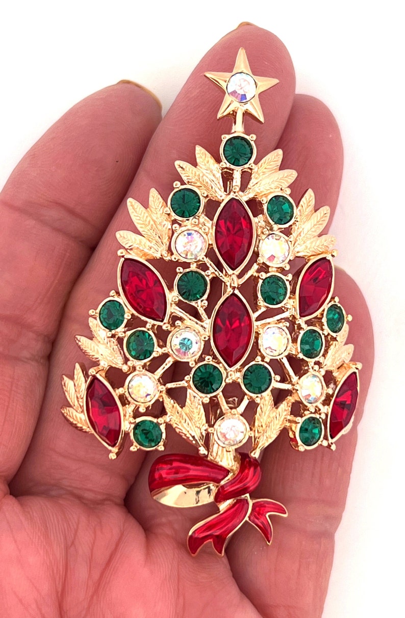 May include: A gold-coloured Christmas tree brooch with red, green, and clear crystal accents. The brooch features a star on top and a red ribbon at the base. The brooch is approximately 6.35 cm tall.