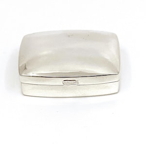 May include: A small, rectangular silver box with a rounded lid. The box has a hinged design and a small clasp on the front. The surface of the box is smooth and reflective, with a bright, polished finish. The box is set against a white background.
