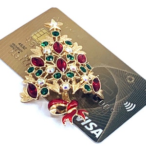 May include: A gold-coloured Christmas tree brooch with red, green, and clear gemstones. The brooch features a red bow and star. The brooch is placed on a gold credit card with the Visa logo.