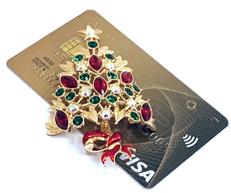 May include: A gold-coloured Christmas tree brooch with red, green, and clear gemstones. The brooch features a red bow and star. The brooch is placed on a gold credit card with the Visa logo.