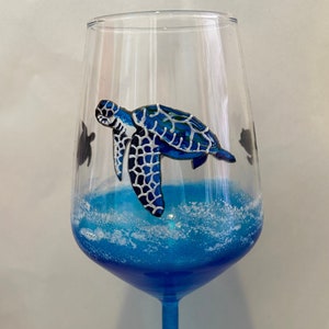May include: A blue glass wine glass with a painted sea turtle design. The turtle is blue, green, and white with a black outline. The glass is filled with blue liquid and white foam.