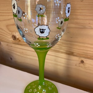 May include: A green wine glass with a white stem and base. The glass is decorated with white sheep and white flowers.
