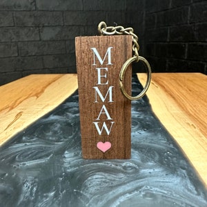 May include: A wooden keychain with the letters "MEMAW" spelled vertically and a pink heart at the bottom. The keychain is attached to a silver chain and a silver ring.
