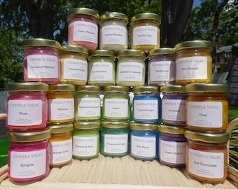 May include: A collection of small glass jar candles with gold lids, arranged on a wooden tray. Each candle has a label with a different scent name, such as "Cherry Blossom," "Rose," and "Hot Chocolate." The candles are filled with colorful wax.