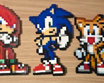 May include: Three pixel art figures made of fused plastic beads. On the left, a red character with white gloves and red shoes. In the center, a blue character with white gloves and red shoes. On the right, a yellow and white character with two tails.